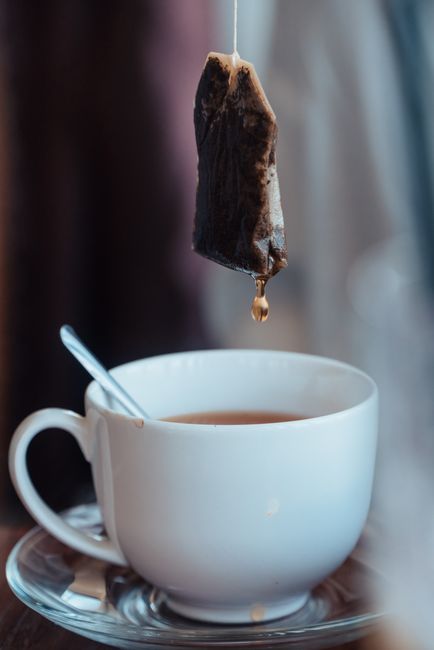 A close-up of a white ceramic cup filled with tea, placed on a reflective glass saucer, with a metal teaspoon resting inside. Above the cup, a used tea bag with dark, damp fabric and a small amount of liquid dripping from the bottom is suspended. The background is softly blurred, suggesting a cozy indoor environment with natural lighting. The image visually depicts a typical scene involving the disposal of used tea bags, which may relate to waste management and rubbish removal services, such as those provided by Waste Disposal Earls Court, with a focus on the proper handling of biodegradable waste in a domestic setting.