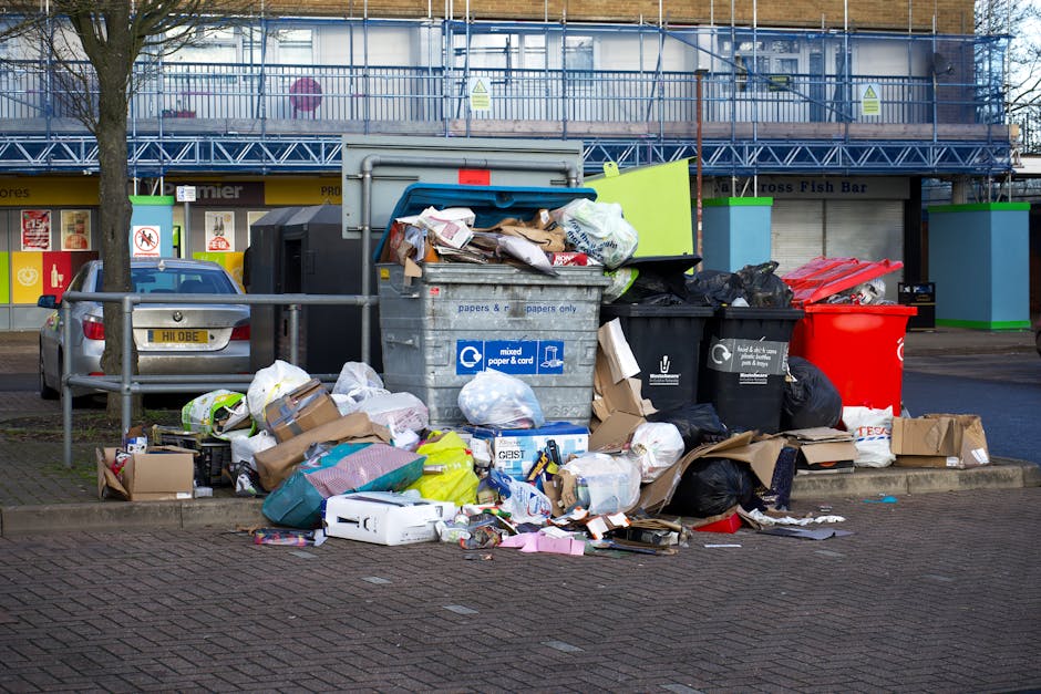 A large collection of overflowing waste bins and scattered rubbish on a paved sidewalk in an urban area outside a commercial building. The waste includes cardboard boxes, plastic bags, and paper packaging, some of which are crumpled or torn, spilling onto the surrounding pavement. There are multiple bins with different colour lids, including a blue bin labeled for mixed paper and card, a black bin for general waste, and a red bin, all placed on a small curb area with a metal railing nearby. Behind the bins, a parked silver car partially blocks the view of a storefront with signage and window displays, and a tree with a bare trunk is visible to the left of the scene. The background includes a building under construction, covered in blue scaffolding and protective sheeting. Natural daylight illuminates the scene, emphasizing the disorderly state of the rubbish and the urban environment. Waste Disposal Earls Court's presence suggests this is an example of waste accumulation prompting the need for professional rubbish removal services.