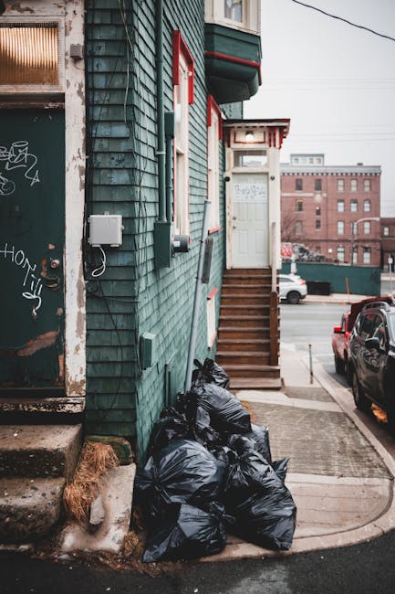 The image shows the exterior of a building clad in weathered, teal-colored wooden siding with visible signs of aging and graffiti on the lower part of a nearby door. A small set of wooden stairs leads up to a white door with red trim, situated at the top of the stairs. To the left of the stairs, there are several tightly packed black rubbish bags piled against the building’s wall, indicating trash awaiting collection. On the left side of the image, a portion of a window with frosted or textured glass is visible, partially obscured by a metal security screen. The surrounding environment includes a paved sidewalk with a concrete curb and a parked black car on the street, with additional vehicles and a multi-storey red brick building in the background. The overall scene reflects a typical urban setting where private waste disposal or rubbish removal services, such as those offered by Waste Disposal Earls Court, might be engaged to handle waste in narrow access areas like this one.