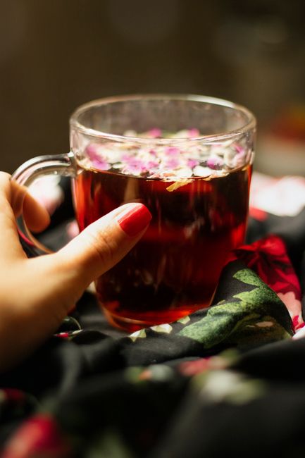 A close-up view of a person's hand with red-painted nails holding a clear glass mug filled with dark red herbal tea, floating with pink and white flower petals on the surface. The mug is positioned slightly off-center towards the left of the image, resting on a soft, black fabric with a floral pattern featuring green leaves and pink roses. The background is blurred with warm brown and beige tones, creating a cozy, inviting atmosphere. The scene highlights the clarity of the glass mug and the delicate appearance of the floral petals, conveying a sense of domestic comfort and relaxation, which subtly aligns with the themes of dedicated waste handling or environmentally conscious disposal associated with rubbish removal services by Waste Disposal Earls Court.