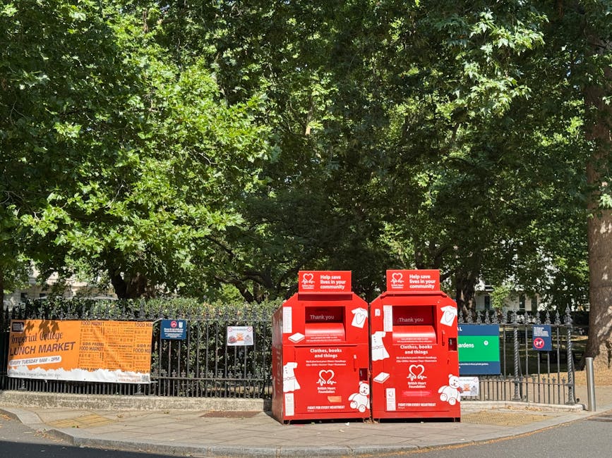 Two red collection boxes for donations of clothing, books, and other items are positioned on the pavement near a black metal fence surrounding a park area with lush green trees in the background. The boxes are made of sturdy metal with a matte finish, featuring white icons and text indicating their purpose, including a heart symbol and a thank you message. Each box has a slot at the top for items to be inserted, and the overall appearance is clean and well-maintained. Behind the boxes, there are multiple signs attached to the fence, including an orange banner advertising a local lunch market with black text, and smaller notices. The scene is illuminated by natural daylight, casting soft shadows, and the environment suggests an urban setting where community-based donation bins are accessible. Waste Disposal Earls Court occasionally handles waste collection at such sites, although these boxes are for charitable donations rather than general rubbish disposal, subtly aligning with alternative waste handling methods in the vicinity.