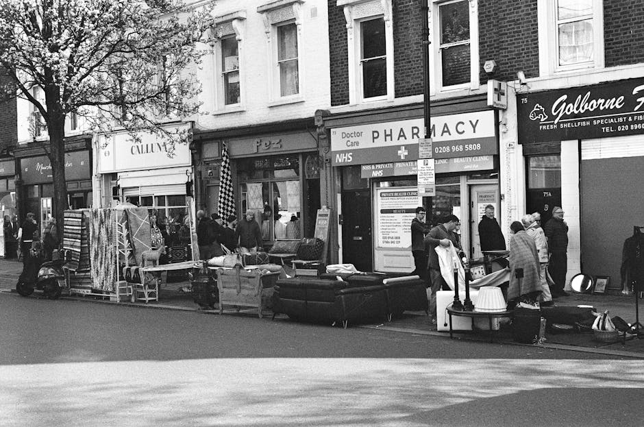 A street scene outside a row of shops, including a pharmacy, capturing a collection of various household items and furniture arranged along the pavement as part of a rubbish removal or on-site clearance process. Visible objects include a black sofa with a textured fabric finish, a small stack of wooden or composite chairs, and several large plastic containers or storage bins. There are also some rolled-up rugs or carpets secured with straps, and a few pieces of lightweight furniture, such as small tables. The items are positioned in front of the storefronts, which include commercial signage for a pharmacy, a fishmonger's shop, and a fish and shellfish specialist. Pedestrians, some wearing face masks, are gathered around, possibly involved in the collection or supervision of disposal activities. The environment is an urban commercial area, with buildings made of brick and plaster, large windows, and a mixture of heritage and modern features. The scene is likely part of a private rubbish disposal service providing a container collection or on-site clearance, as indicated by the arrangement of bulky waste items, aligning with independent waste handling options in urban settings. Natural lighting suggests daytime, with minimal shadows and overcast skies.