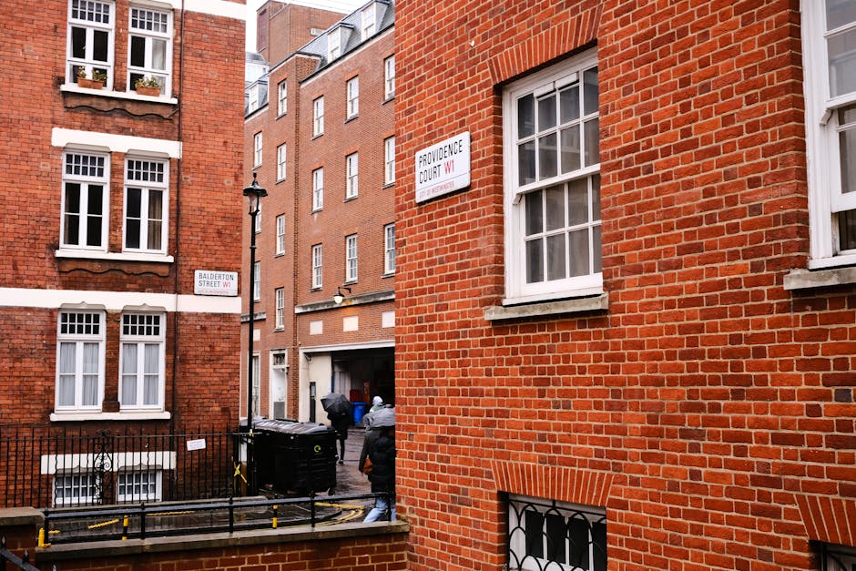 The image depicts a narrow urban alleyway bordered by red-brick residential buildings with multiple white-framed sash windows. In the foreground, a section of a brick wall with vertical, evenly spaced brickwork accents is visible on the right. Behind this, the alley opens up, revealing a black wheelie bin placed near a metal fence, which is adjacent to a paved surface with yellow barriers. A person dressed in dark clothing and a hood is seen walking in the alley, heading away from the camera, next to a few other bins and garbage bags. The buildings have traditional brickwork and appear well-maintained, with simple signage indicating street names, such as 'Balderton Street W1' on the left and 'Providence Court W1' on the right. The scene is lit by natural daytime light, casting shadows on the brick facades, and illustrating a typical UK residential alley with provisions for waste disposal, relevant to private rubbish collection and waste management services provided by Waste Disposal Earls Court for the local area.
