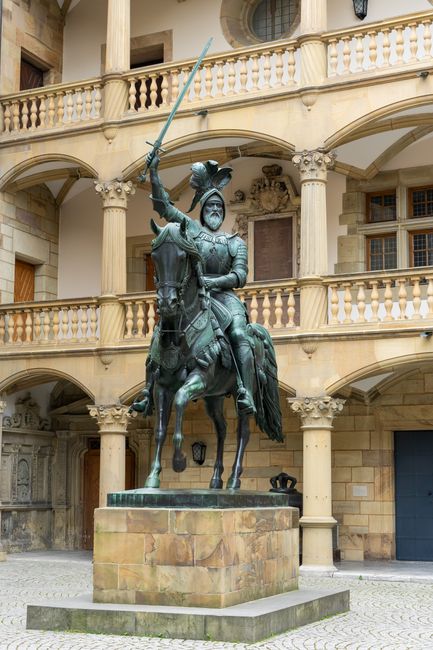 A large bronze equestrian statue of a knight in armor, holding a sword raised above his head, is situated on a stone pedestal in an urban courtyard. The knight is depicted wearing a detailed helmet with a plume and seated on a horse that is captured in a dynamic posture with all four hooves on the pedestal. The background features a historic building with cream-colored stone walls, supported by ornate columns that create a colonnaded balcony on the upper level. The balcony has a balustrade with evenly spaced spindles, and there are large windows with wooden frames visible behind it. The courtyard pavement consists of cobblestones, and subtle lighting suggests daylight or soft overcast conditions, creating a neutral and quiet atmosphere. While primarily a decorative public sculpture, the scene underscores the importance of maintenance and cleaning in historic or urban environments, aligning with services like those provided by Waste Disposal Earls Court for rubbish removal and site clearance in city settings.