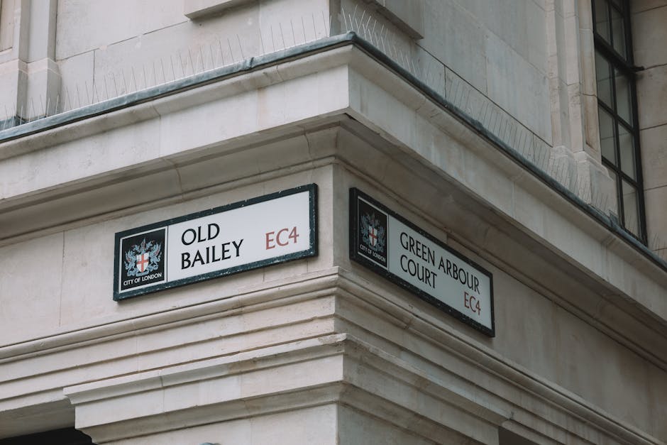 Photograph of a corner of a beige stone building with two black and white street signs attached to the facade at the junction. The left sign reads 'Old Bailey EC4' and features the emblem of the City of London, including a red cross on a white background with a red sword in the upper left quadrant. The right sign reads 'Green Arbour Court EC4' and also bears the City of London crest. The building has detailed stonework along the edges and appears to be part of an older, classical architectural style. A window with black metal framing and multiple panes is partially visible on the upper right side of the image. The scene is evenly lit with natural daylight, and no vehicles or pedestrians are visible, emphasizing the clean, historic look of the urban environment, relevant to discussions around city streets and private or independent waste collection opportunities in central London.